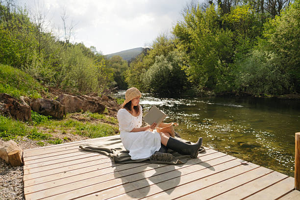 Photo of a young woman reading, soaking the sun and relaxing on a riverside dock
