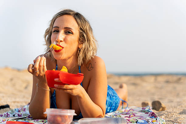 Woman lying on beach towel eating fresh fruit salad from reusable container