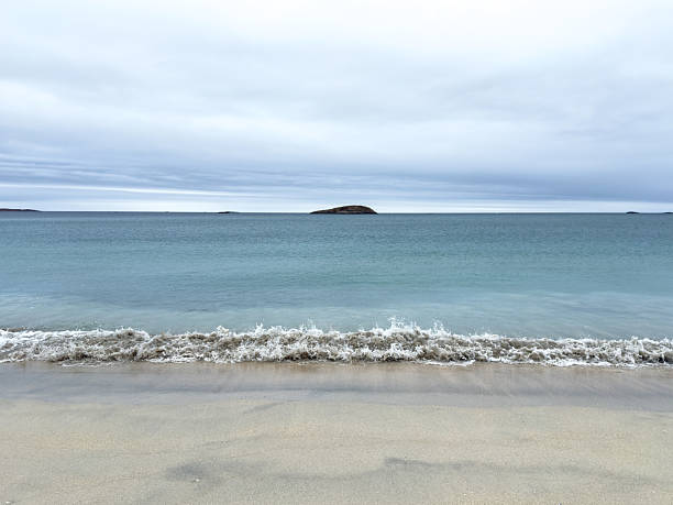 Serene shoreline with soft waves lapping at sandy beach. Distant island under a cloudy sky. Calm ocean scene for relaxation and travel themes.
