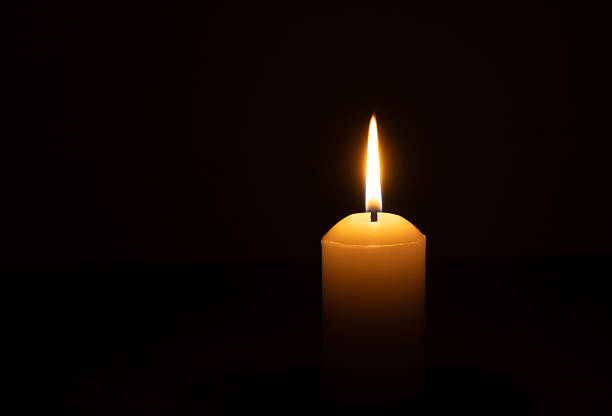 istock A single burning candle flame or light glowing on a big white candle isolated on black or dark background on table in church for Christmas, funeral or memorial service 2218783367