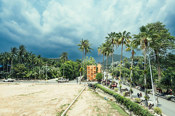 Chon Buri, Thailand - May 1, 2023 - The sky is cloudy and the sun is hidden behind the clouds. There are palm trees in the background and a building in the distance. The scene is peaceful and calm, with people walking around