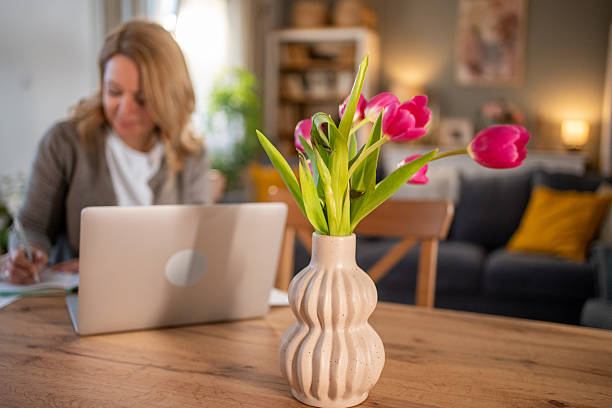 Bright pink tulips in a decorative vase on a wooden table enhance a cozy home office, while a mid-adult woman works on a laptop in a warm, productive atmosphere