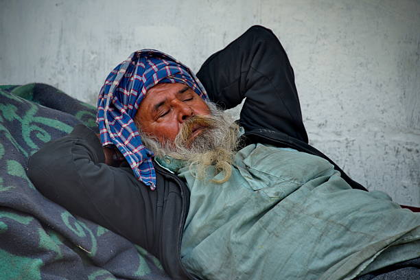 Middle aged Black man lying on makeshift bed inside tent holding hands together, wearing winter clothing and scarf, suitcase placed behind head, resting on pillow on ground