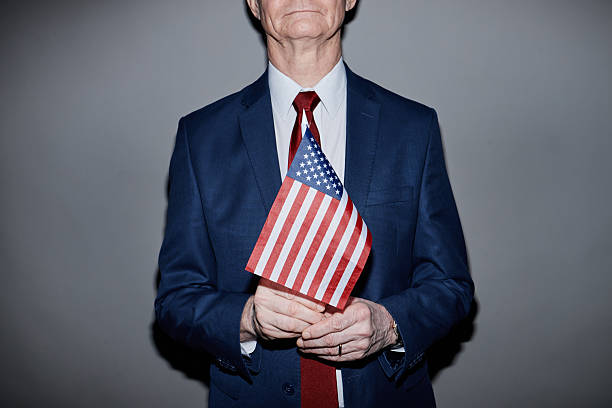 Cropped shot of Caucasian senior male political candidate in suit and red tie holding American flag while standing against grey background in studio, camera flash