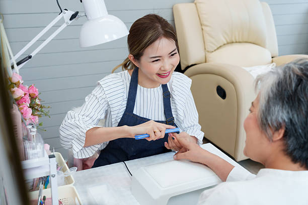 istock Young nail spa technician smiling while filing senior asian woman's nails during cozy manicure session promoting beauty personal grooming hygiene wellness relaxation lifestyle in small business salon 2218792503