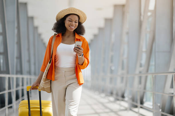 istock Black woman walking through airport terminal, checking phone and pulling yellow suitcase. 2218792615