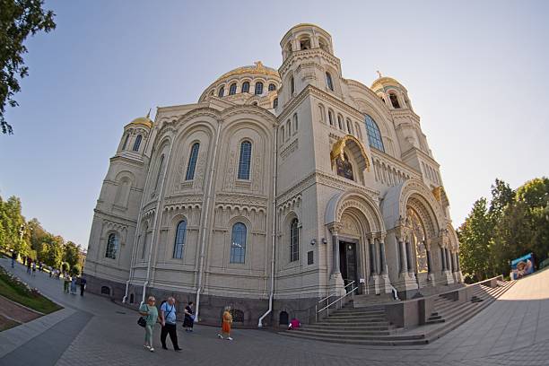 Kronstadt, Russia, September 5, 2024.Naval Cathedral of Saint Nicholas in Kronstadt, Russia, captured with a wide-angle lens. The grand structure features a light stone facade, ornate arches, and multiple domes. Intricate architectural details include arched windows and decorative moldings. The cathedral s vertical stature dominates the scene, set against a clear sky. Steps lead to the entrance, and people, appearing as small figures in comparison, walk nearby. Trees and manicured landscaping frame the foreground, set on a stone platform.