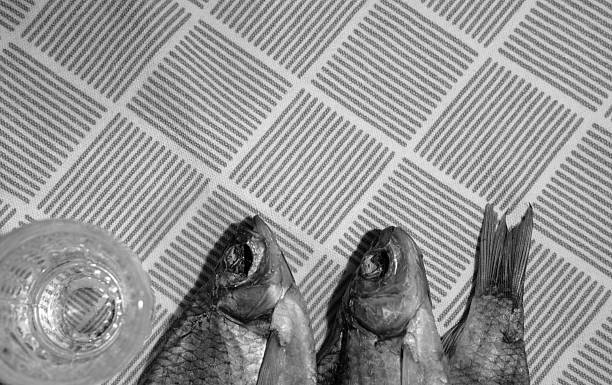 Minimalist close-up image of two dried fish and a traditional vodka glass placed on a light fabric with a geometric pattern. The setup reflects a culturally rich, rustic eating experience typical in Slavic and Baltic countries. Ideal for illustrating food traditions, cultural heritage, and minimalist still life scenes.