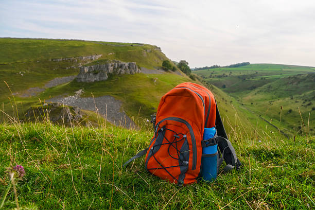 A bright orange backpack sits on lush green grass at the edge of a hill, showcasing a stunning valley with rolling green hills under a clear sky.