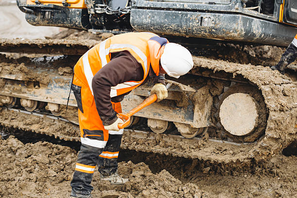 Construction worker  in bright safety gear working hard using shovel to clean tracks of his heavy machinery excavator on muddy site