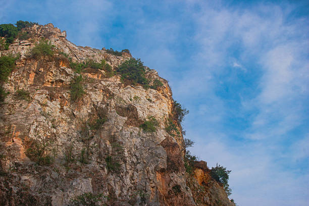 A close-up view of a mountain peak captures natural rock textures , cracks , and weathering. Sparse green foliage clings to the rugged surface beneath a clear blue sky with light , scattered clouds.
