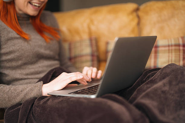 Young woman with red hair using laptop while sitting on a cozy couch wrapped in a blanket during a relaxed indoor moment