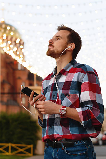 Young man enjoying music outdoors under city festive lights. Guy with beard and modern hairstyle wearing in blue, red and white plaid shirt walking through the city streets in urban background.