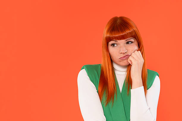 istock Red-haired woman with a thoughtful expression against an orange background while wearing a green vest and white sweater 2218869100