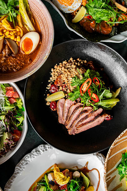 A variety of gourmet dishes are beautifully presented, including a bowl of ramen with a soft-boiled egg, a fresh salad with greens and tomatoes, and sliced meat garnished with herbs, nuts, and pickles
