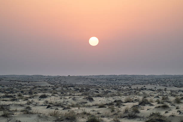 A captivating image captures the dark sunset over the vast desert landscape of Dubai. The sky glows with deep hues of orange, red, and fading gold as the sun dips below the horizon, casting long shadows across the rolling sand dunes. Silhouettes and subtle textures emerge under the twilight, creating a dramatic and serene atmosphere. The remote, arid environment evokes a sense of solitude and mystery, blending natural beauty with the quiet stillness of dusk in the wilderness