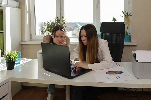 istock Two female coworkers at a shared workspace reviewing printed materials and collaborating digitally on a laptop, embodying concepts of office synergy, project management, and productive collaboration. 2218893393