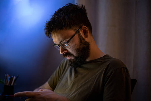 A man using a digital device in the dark at night, illuminated by screen light, sitting alone in his modern apartment.