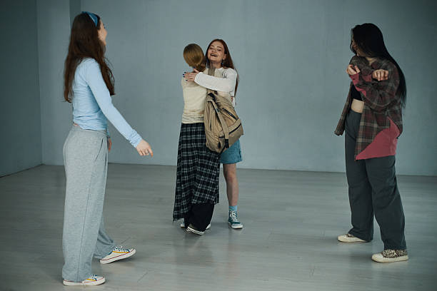 Group of four young women interacting and hugging indoors against a plain backdrop, with two standing and two embracing, creating an uplifting atmosphere