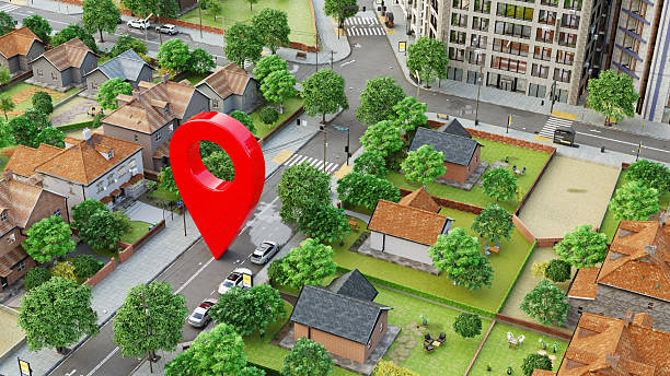 Digitally rendered illustration of a suburban neighborhood with houses, trees, and streets. The central focus is a large, red map location pin marker hovering over a street, symbolizing a geolocation or navigation point - commonly associated with GPS, maps, or location-based services. The setting is clean and modern, with a mix of single-family homes and urban buildings in the background, emphasizing the integration of digital tools with real-world environments.
