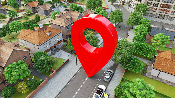 Digitally rendered illustration of a suburban neighborhood with houses, trees, and streets. The central focus is a large, red map location pin marker hovering over a street, symbolizing a geolocation or navigation point - commonly associated with GPS, maps, or location-based services. The setting is clean and modern, with a mix of single-family homes and urban buildings in the background, emphasizing the integration of digital tools with real-world environments.