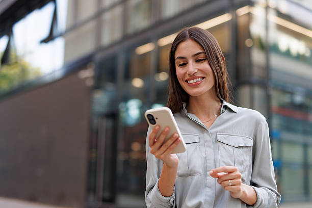 Professional female professional networking on mobile device, standing near contemporary skyscrapers with confident smile