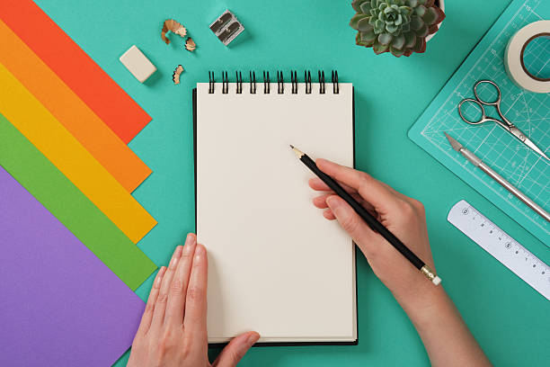 A person is preparing to draw on a spiral-bound notebook with a black pencil, surrounded by colorful sheets of paper and craft tools. Space for your text or drawing