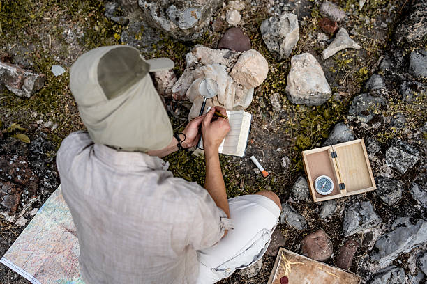 Top-view of a person outdoors exploring with a map, compass, notepad, and magnifying glass among stones.