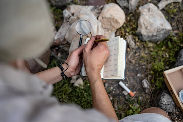A person sits outdoors holding a notebook, writing with a pen, with a magnifying glass and other tools placed on the ground nearby, amidst rocks and greenery, suggesting exploration or research activities.