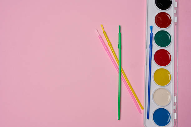 A set of watercolor paints in a plastic case with multi-colored brushes is located on a pink background.