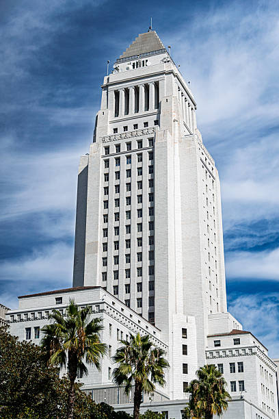 Low-angle view of the iconic Los Angeles City Hall, a historic government building with Art Deco architecture, standing tall under a vivid sky. Palm trees frame the base, adding a Southern California feel.