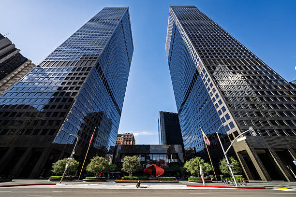 Contemporary office buildings and glass skyscrapers tower above the streets of downtown Los Angeles on a bright, sunny day. The sleek architecture, clean sidewalks, and reflections of the blue sky highlight the city's modern urban character and business district atmosphere.