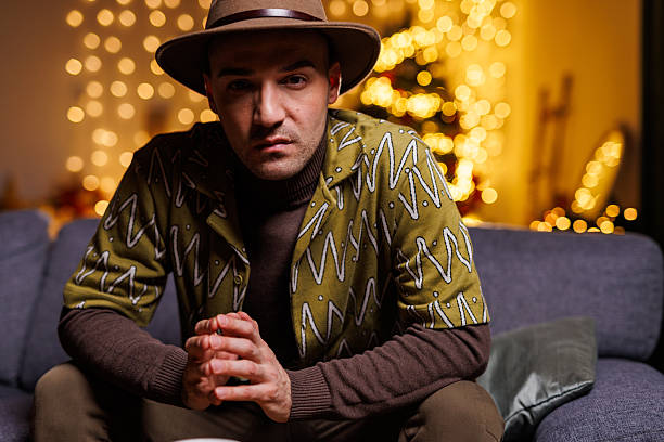 A stylish man wearing a patterned shirt and hat sits on a couch indoors, surrounded by warm holiday lighting, creating a cozy and festive ambiance with a touch of modern fashion and personality.