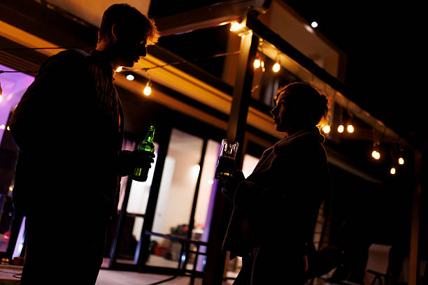 Two individuals enjoy beverages while standing in front of a modern house decorated with string lights at nighttime, creating a relaxing and calming atmosphere.