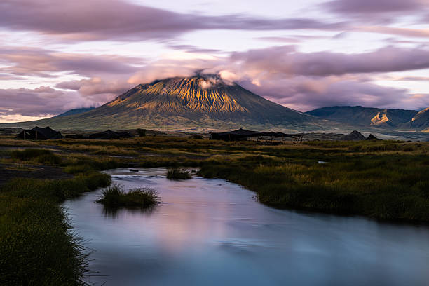 Soft dawn light grazes the dramatic slopes of Ol Doinyo Lengai, the sacred volcano towering over the plains near Lake Natron, Tanzania. A slow-flowing stream in the foreground reflects the purples and golds of the sky, while scattered Maasai shelters rest under the volcano’s shadow—capturing the mystic beauty of this remote East African landscape.