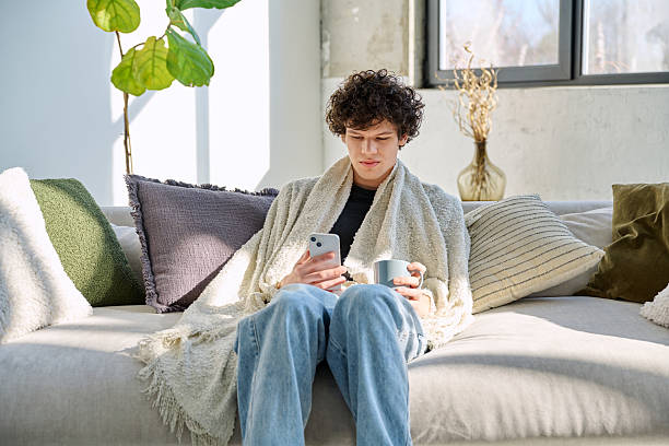 istock Young relaxed guy under warm blanket with mug smartphone, sitting o sofa at home 2218995583