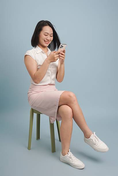 istock Woman using a smartphone while sitting on a green stool against a light blue background in a casual setting 2219006248