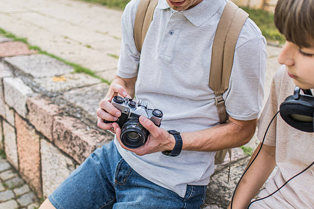 Old skills versus new technology. Father teaching his son how to use retro photo camera and how to take pictures while sitting near the scenic urban path along the riverbank.
