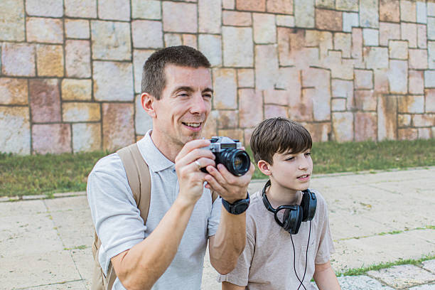 istock Old skills versus new technology. Father teaching his son how to use retro photo camera and how to take pictures while sitting near the scenic urban path along the riverbank. 2219009852