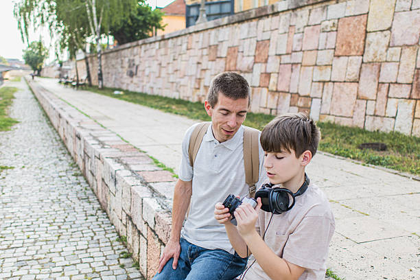 istock Old skills versus new technology. Father teaching his son how to use retro photo camera and how to take pictures while sitting near the scenic urban path along the riverbank. 2219009973