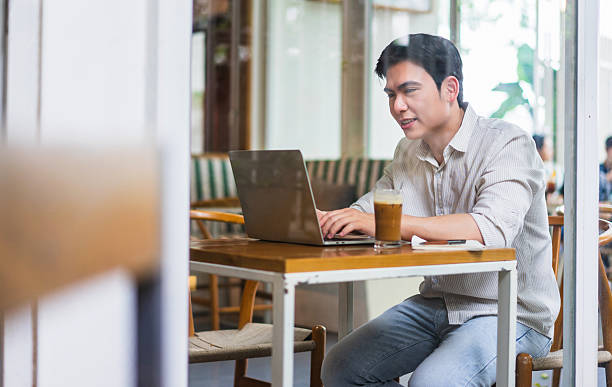 istock Young Asian man working on laptop in a modern cafe during the afternoon while enjoying iced beverage 2219011814
