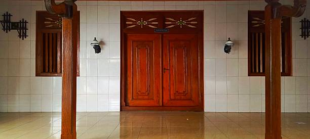 This image shows a symmetrical architectural detail with a central wooden double door, flanked by two windows of similar design. The walls are tiled in white, and the floor is a light-colored tile, reflecting some light. Two wall-mounted lamps are visible on either side of the door, and there are two sturdy wooden pillars in the foreground. The overall impression is one of traditional craftsmanship and calm.