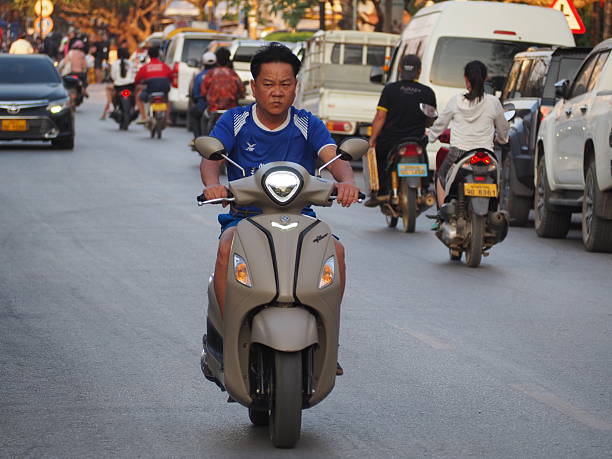 Luang Prabang, Laos – February 19, 2025: People ride scooters and motorbikes through the streets of Luang Prabang, a UNESCO World Heritage city known for its mix of traditional Lao architecture and colonial influences. The scene reflects everyday transportation in Laos, with scooters weaving through narrow roads, passing by shops, trees, and local homes. It captures the rhythm of daily life in this historic and culturally rich Southeast Asian city.