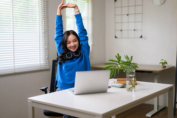 istock Happy young Asian woman stretching her arms while enjoying her work-from-home routine, sitting comfortably at her desk with a laptop 2219021379