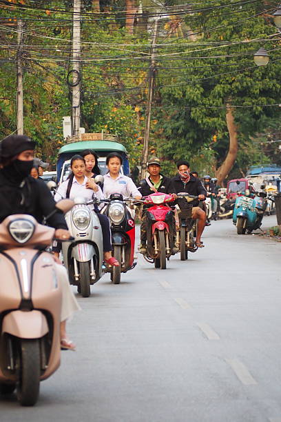 Luang Prabang, Laos – February 19, 2025: People ride scooters and motorbikes through the streets of Luang Prabang, a UNESCO World Heritage city known for its mix of traditional Lao architecture and colonial influences. The scene reflects everyday transportation in Laos, with scooters weaving through narrow roads, passing by shops, trees, and local homes. It captures the rhythm of daily life in this historic and culturally rich Southeast Asian city.