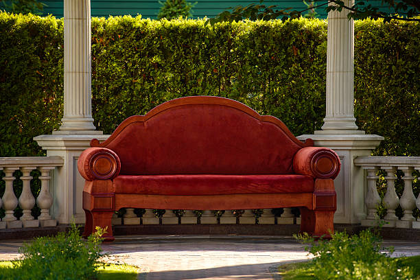 Luxurious red vintage-style sofa with wooden details placed in a classical garden pavilion. Surrounded by white columns, balustrades, and greenery. Perfect setting for romantic or elegant scenes.
