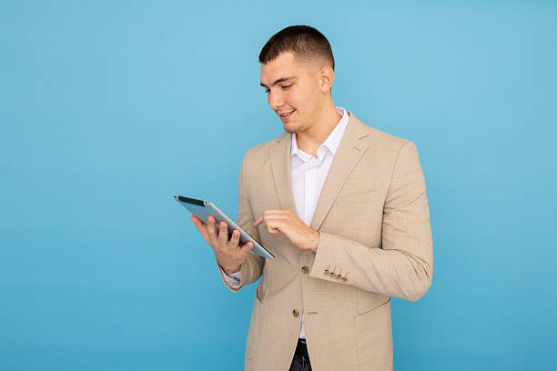 istock Young adult businessman smiling while using a tablet, scrolling and tapping against a vibrant blue background in a studio setting 2219029381