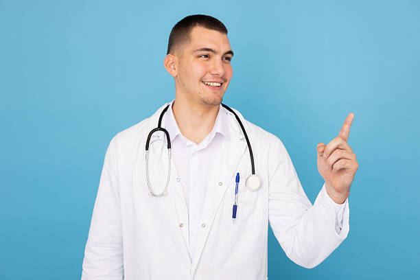 istock Medical professional wearing white lab coat and stethoscope, pointing confidently against bright blue background 2219030129