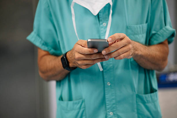 istock Doctor in surgical scrubs using smartphone in hospital hallway, focused on screen while holding phone with both hands 2219035440
