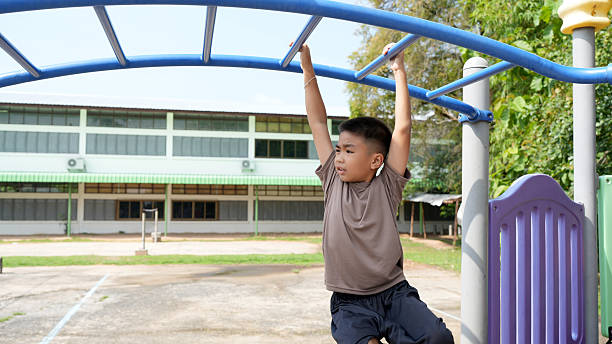 istock Young boy is playing on playground, hanging from blue monkey bar with school building in background. scene captures moment of joy and physical activity in sunny environment 2219043976